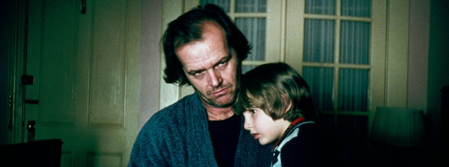 Jack Torrence (Jack Nicholson) sits on a bed, holding his son Danny who is sitting on his lap