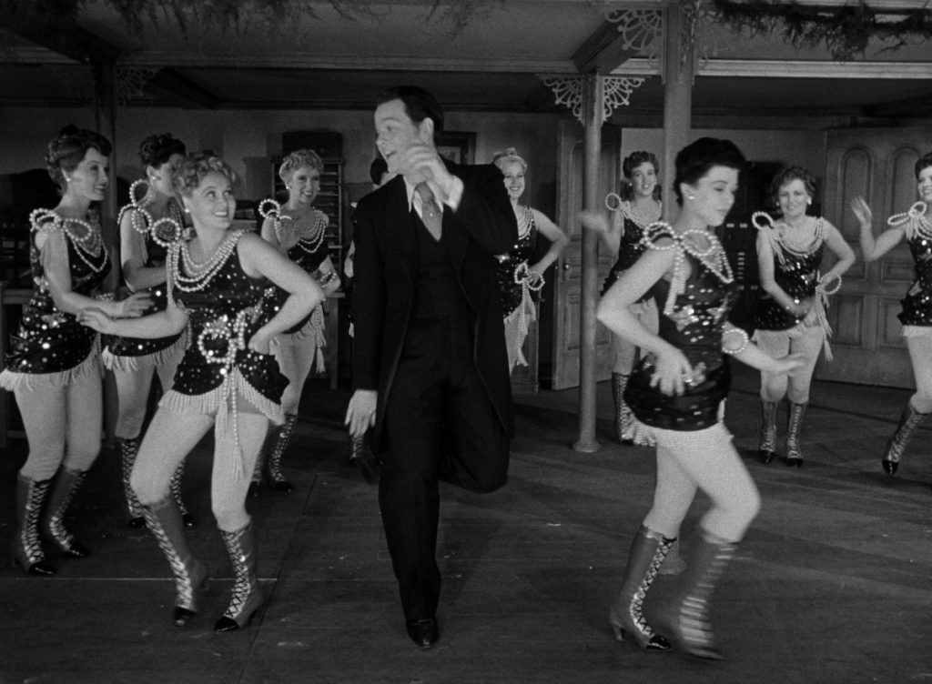 Orson Welles as Charles Foster Kane in a business suit dances with a troupe of chorus girls in sequin and pearl dresses