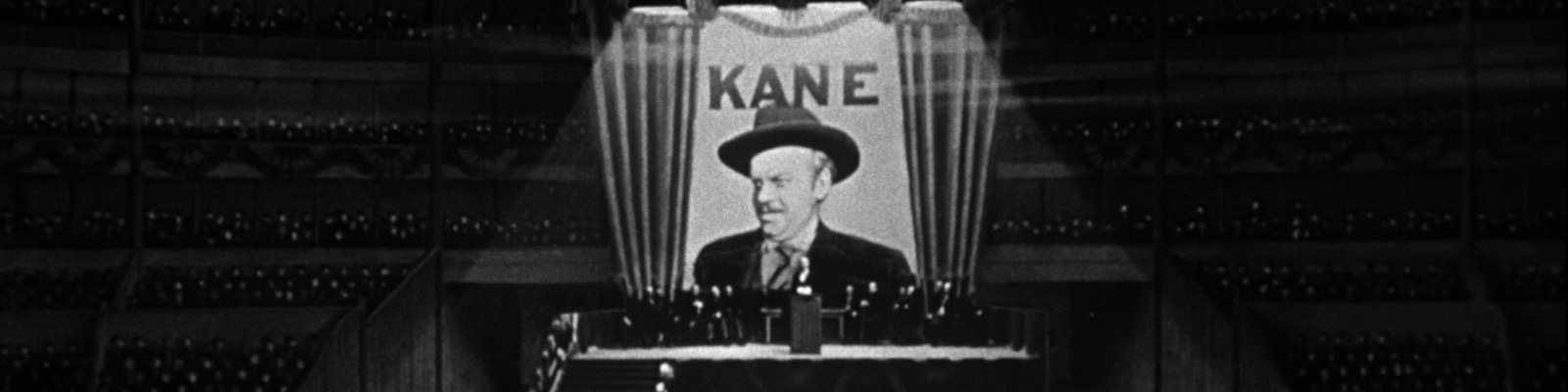 Orson Welles as Charles Foster Kane stands on stage in front of a large poster bearing his image and the word KANE