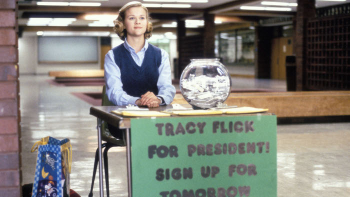 Reese Witherspoon as Tracy Flick in Election sits primly at a desk in a high school. Attached to the front of the desk is a handmade sign reading "Tracy Flick for President! Sign up for tomorrow." On the desk are a glass fishbowl filled with election buttons and three clipboards. 