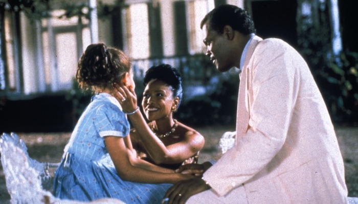 Jurnee Smollett wearing a prim dress sits outside a stately home on an ornate white painted iron bench. Samuel L Jackson in a white suit sits across from her holding her hand, and Lynn Whitfield wearing a strapless evening gown kneels while caressing Jurnee's face.