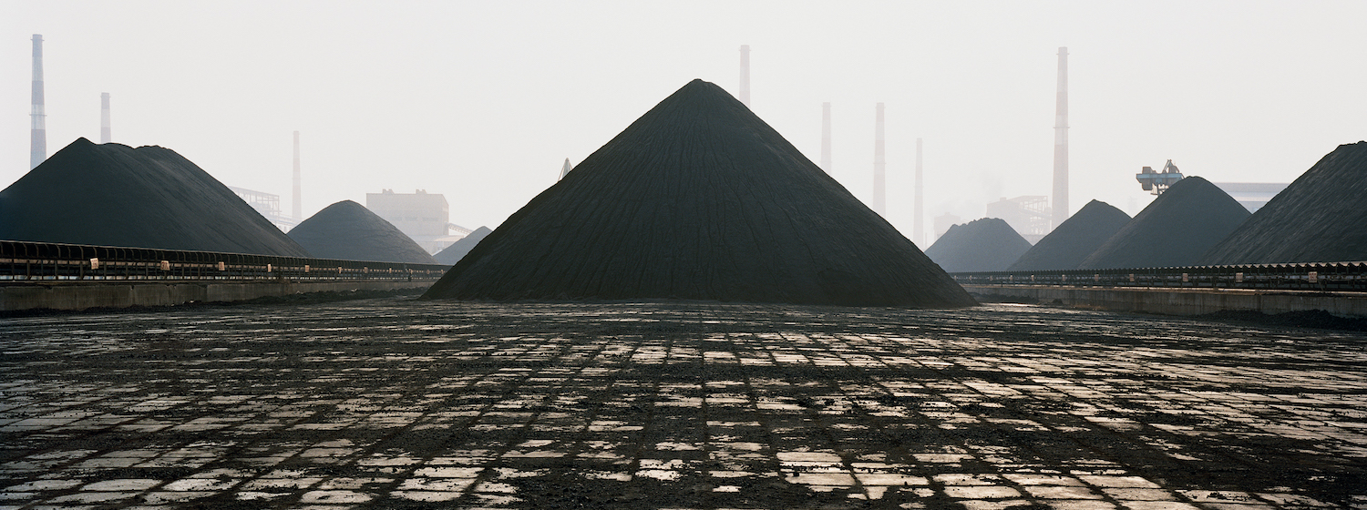 A shot of a polluted industrial site featuring enormous coal heaps and many smokestacks under smoggy skies in Manufactured Landscapes