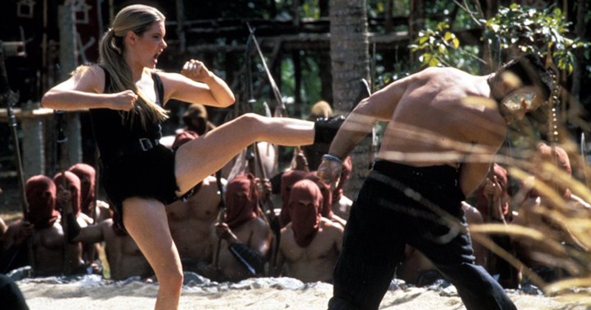 Bridgette Wilson performing as Sonya Blade in Mortal Kombat (1995) kicks Trevor Goddard appearing as Kano in a fight scene in a jungle surrounded by masked figures.