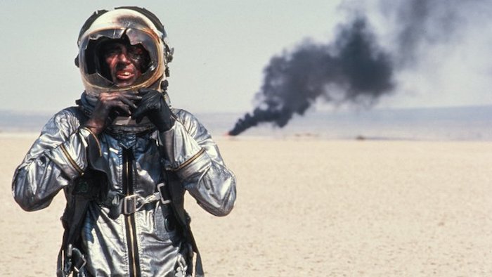 A pilot in a space suit with a smashed face shield stands in a desert. Black smoke is rising in the distance. Scene from The Right Stuff.