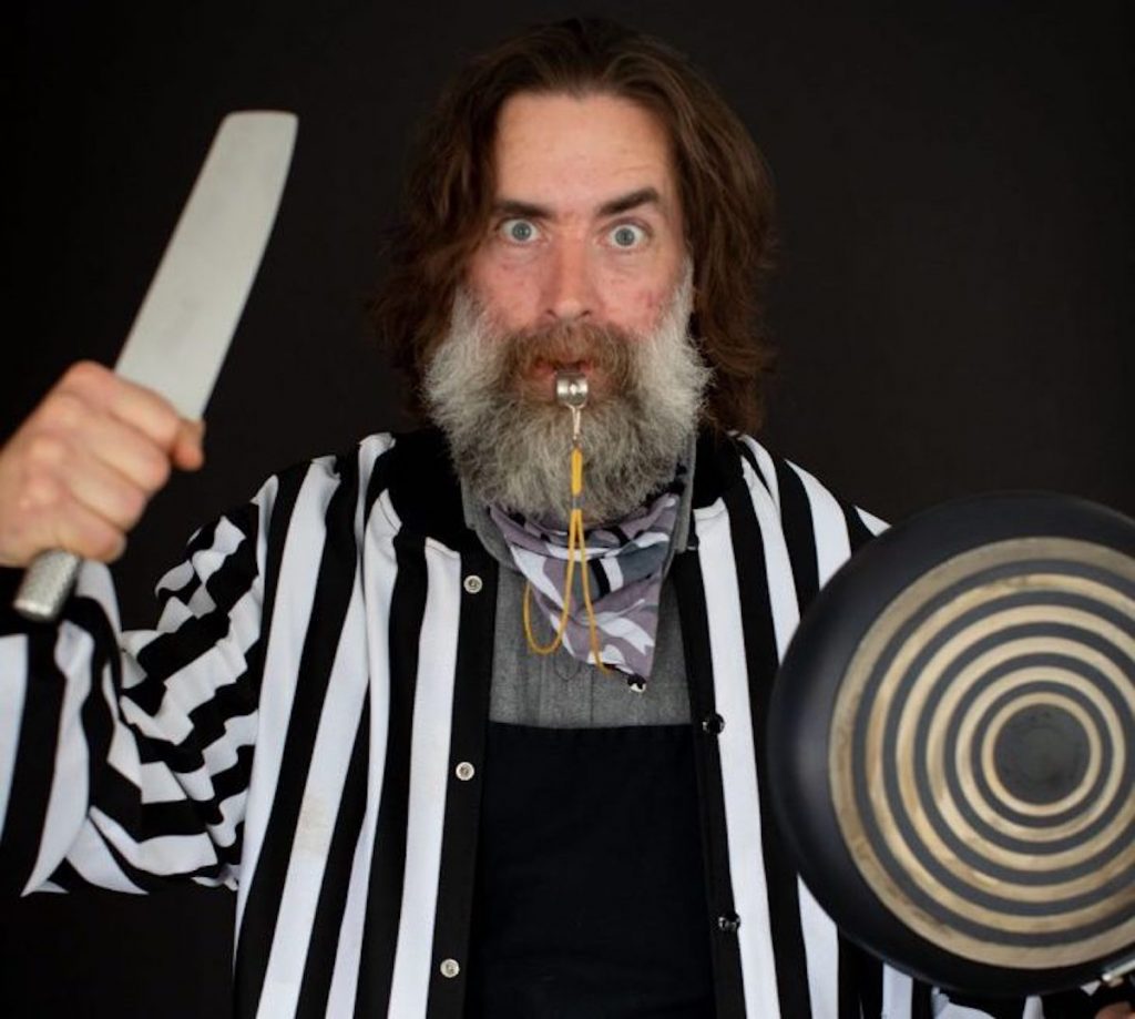 Sam Higgs poses for a portrait wielding a cooking knife and a frying pan.