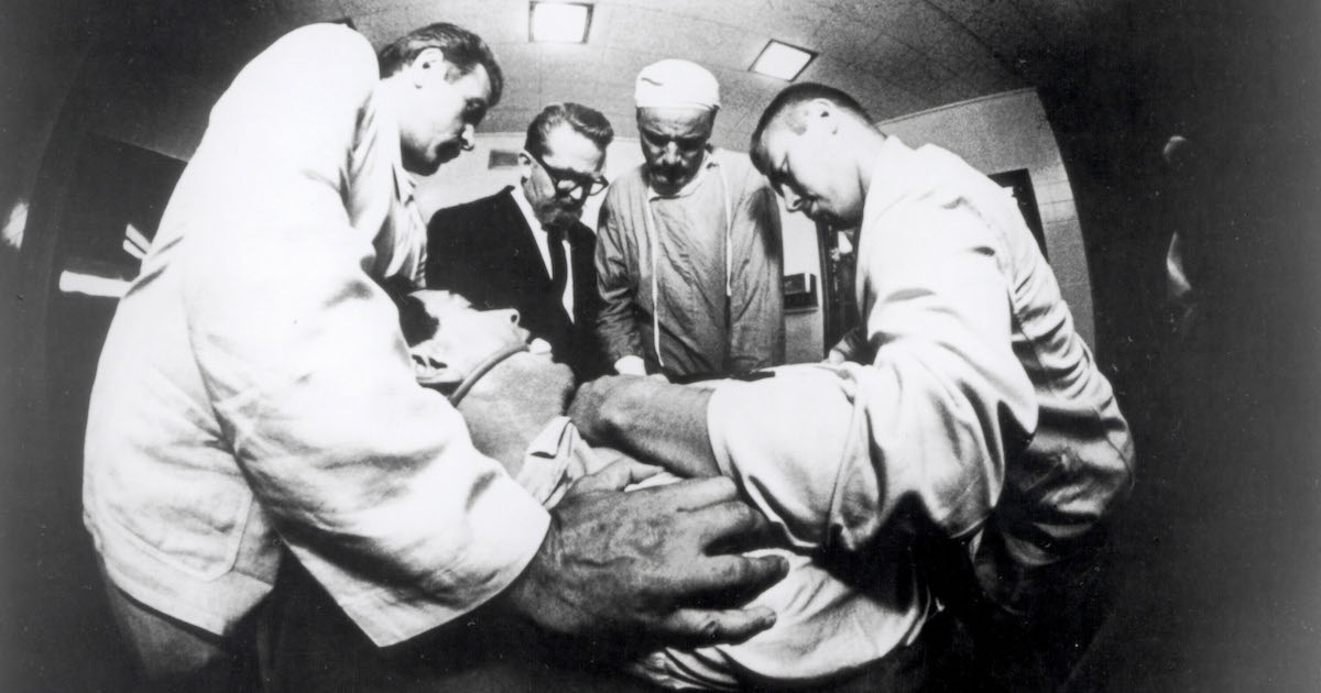 People in white coats and medical scrubs surround Rock Hudson who is laying back in a chair