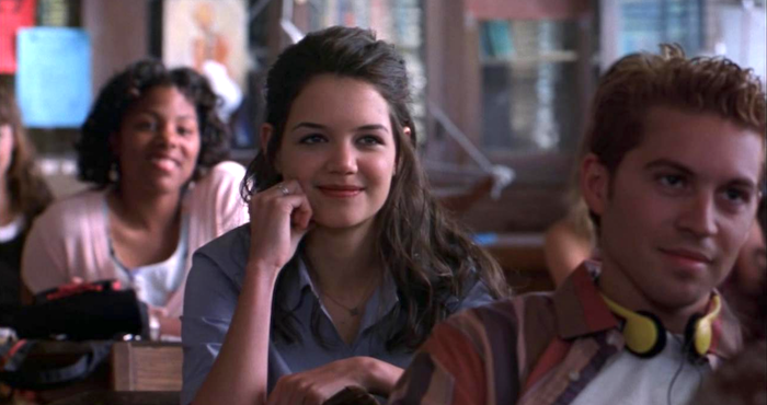 Katie Holmes, smiling sits at a desk in a classroom surrounded by other students
