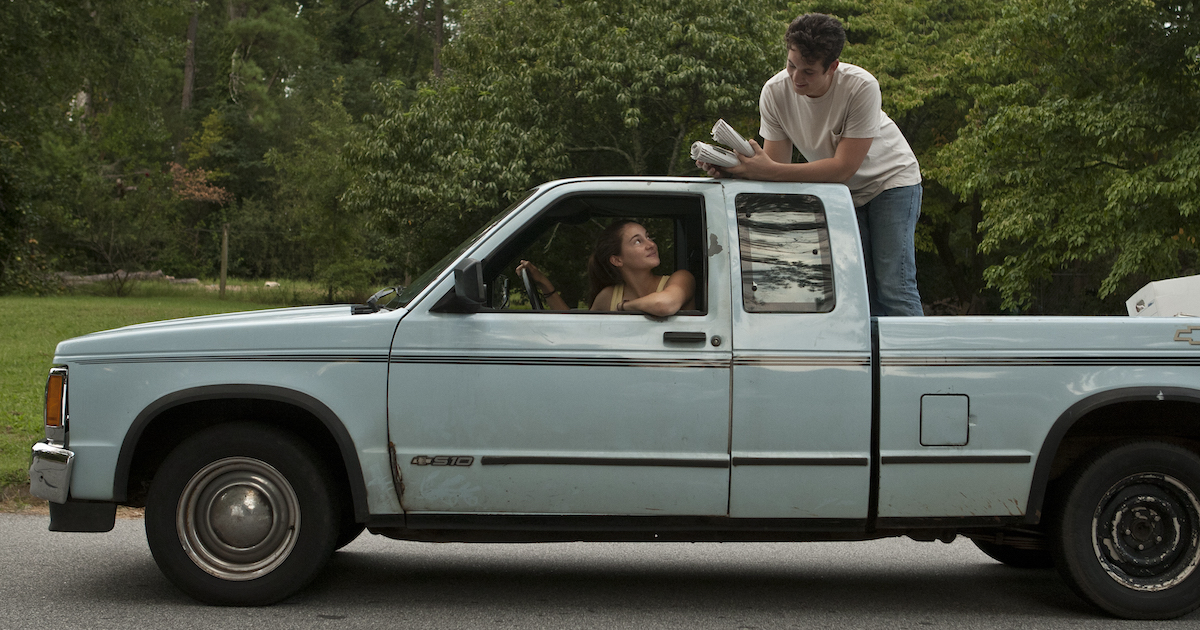 Shailene Woodley drives a pickup truck as Miles Tellar stands in the back holding newspapers he is delivering in The Spectacular Now