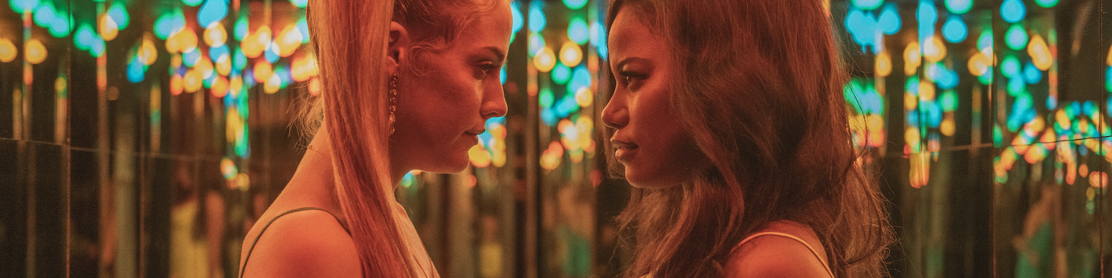 Riley Keough and Taylour Paige stand face to face in a small mirrored corridor with bright lights lining the ceiling. Image capture from Zola