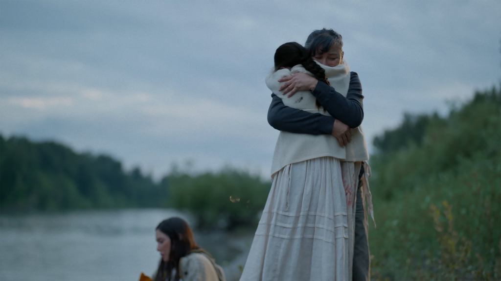 Two women hug as a third sits nearby along the forested shore of a body of water. Still from the film Angela's Shadow