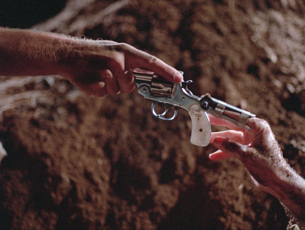 in a dirt field, a close up of a pistol being passed between two hands in reference to Michaelangelo's "The Creation of Adam" fresco in the Sistine Chapel