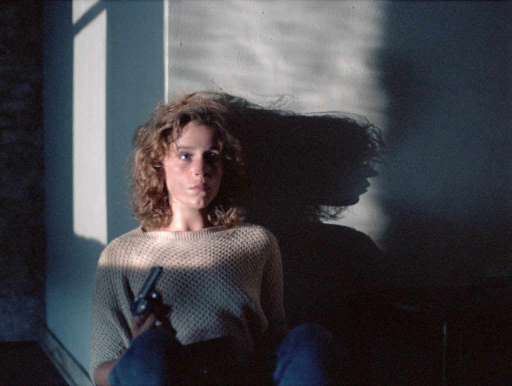 Looking apprehensive, Frances Mcdormand sits leaning against a wall holding a pistol