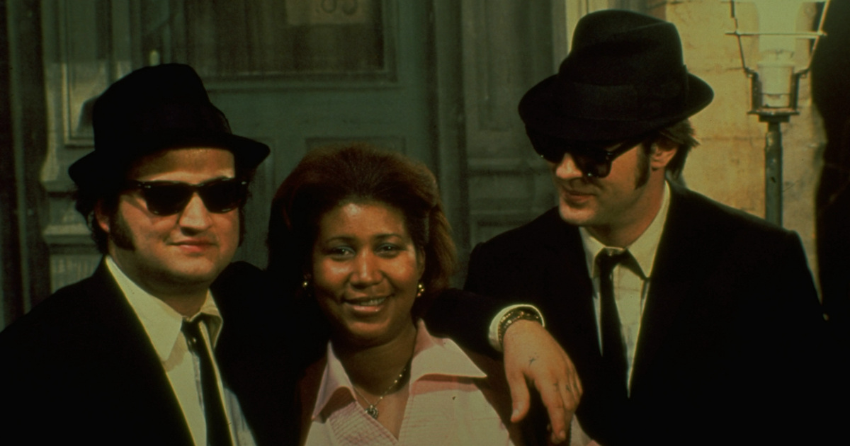 John Belushi and Dan Aykroyd in black suits, fedoras and ray ban sunglasses appear as the Blues Brothers in a photo with Arthea Franklin in the film The Blues Brothers