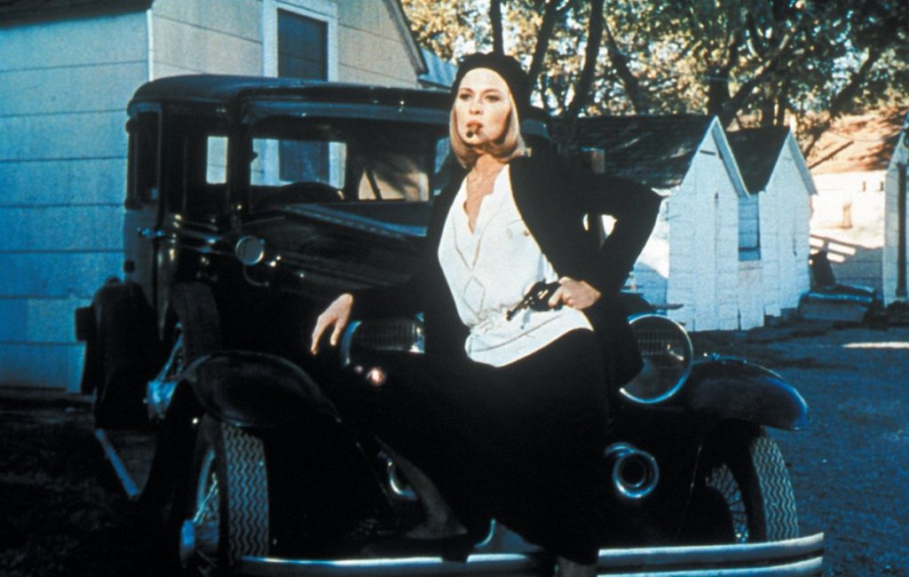 Bonnie (Faye Dunaway) poses in front of a car with a pistol at her hip and a cigar in her mouth