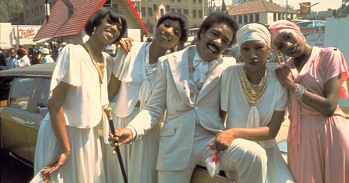Richard Pryor, wearing a white suit with a large ruffled bow tie, and an oversized, jewelled cross on a thick gold chain holds a cane as he sits on the hood of a golden limosine surrounded by young, well dressed women in Car Wash.