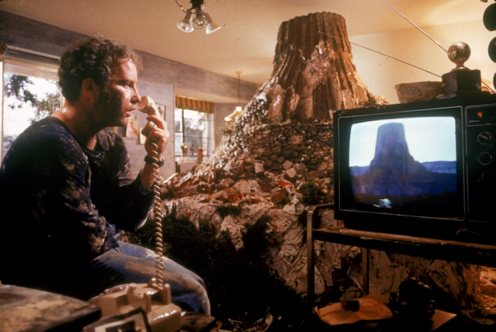 Richard Dreyfus holds a rotary telephone while looking at TV showing the Devil's Tower rock formation. Behind the TV is a large model of Devil's Tower Dreyfus' character has built in his home.
