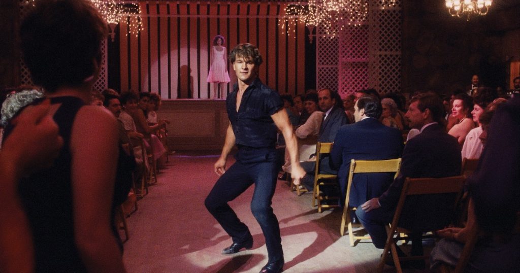 Patrick Swayze and Jennifer Grey dance in the final scene of in Dirty Dancing. Swayze dances in the aisle of an auditorium as Grey stands on the stage in the background