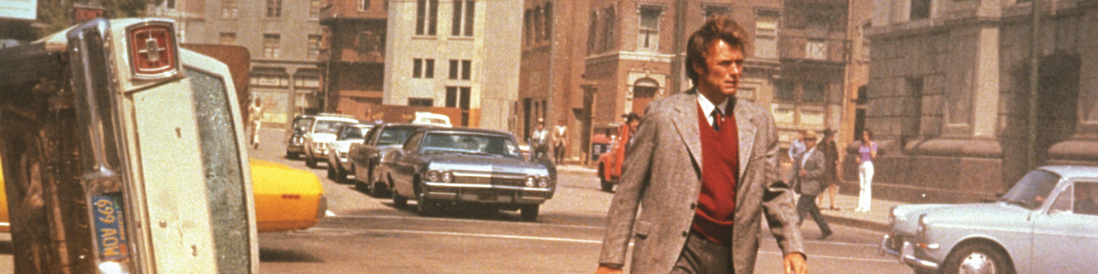 Clint Eastwood holding a gun walks down a street beside a car flipped on its side in Dirty Harry