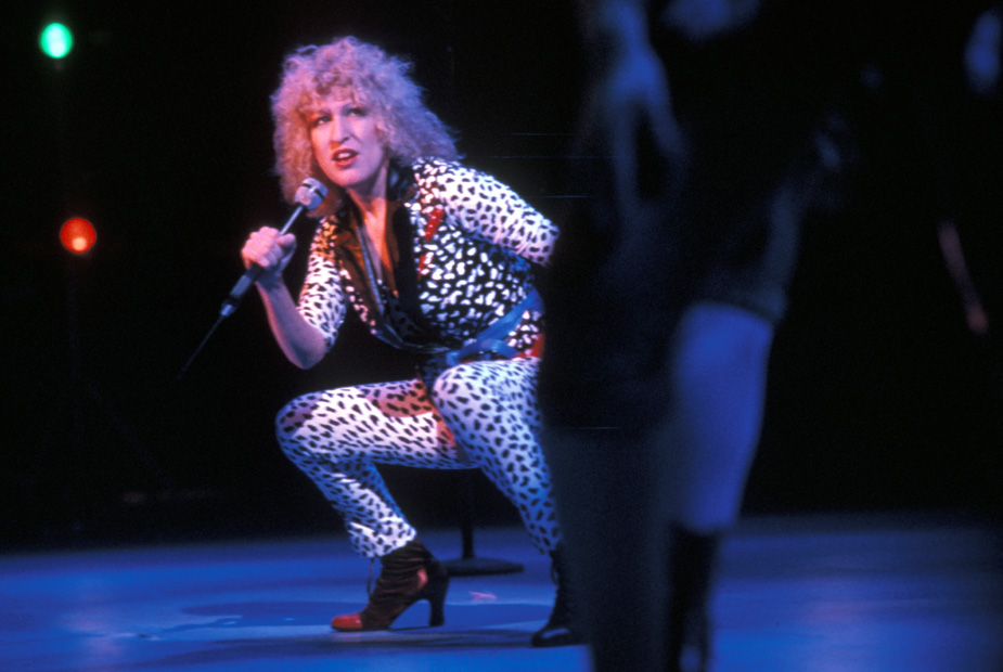 Bette Midler, wearing a black and white spotted jump suit, crouches a she performs in the concert film Divine Madness