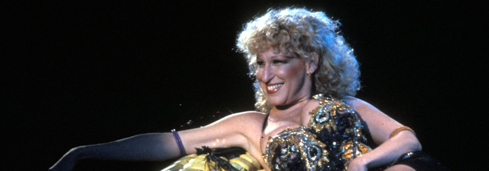 Bette Midler, wearing a a sequin and gem covered bodice, smiles as she leans against an enormous prop ear of corn on stage in the concert film Divine Madness