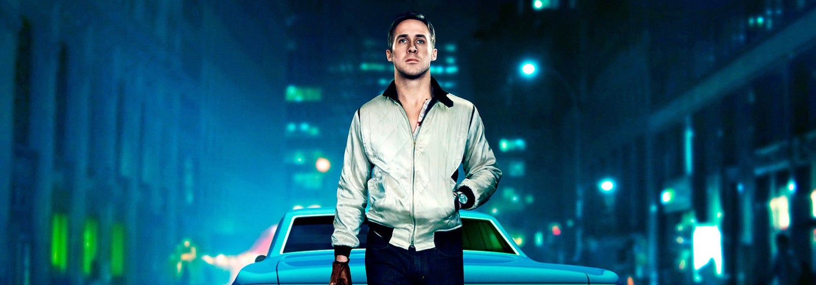 Ryan Gosling stands behind a car on a blue-lit city street in Drive