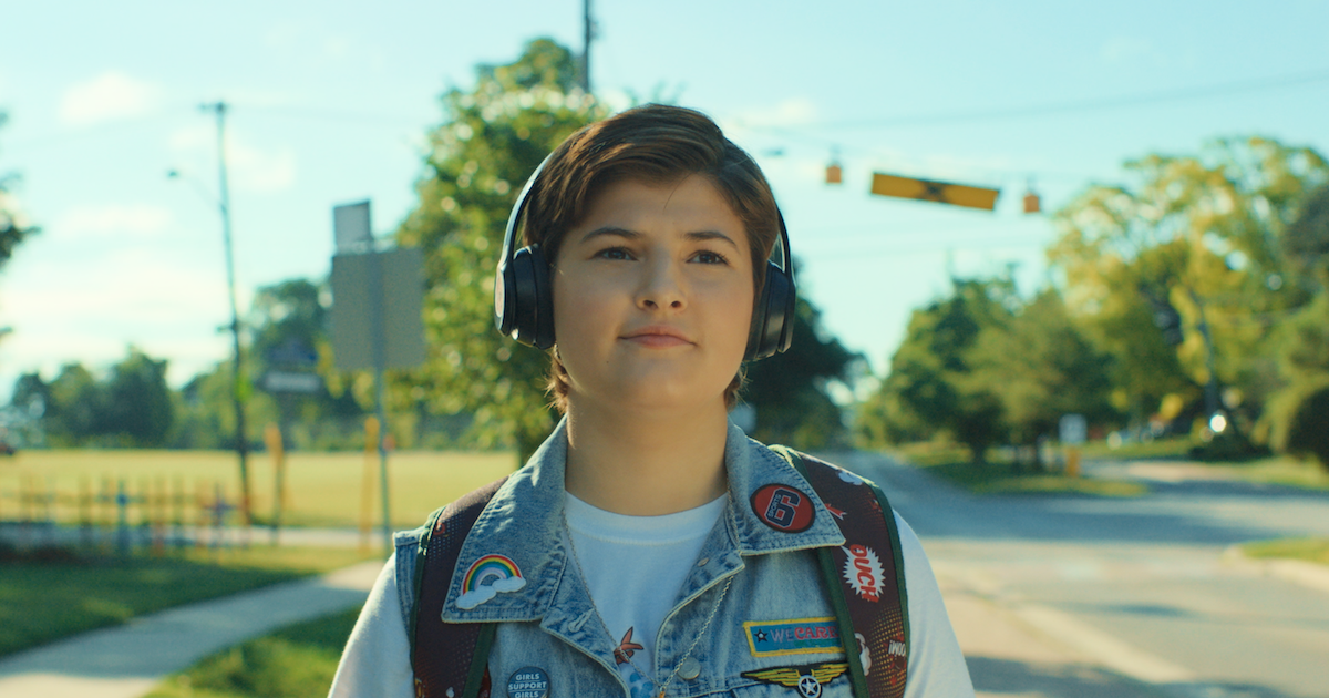 Elliot Stocking (Erin in the film Erin's Guide to Kissing Girls) is a white teenager with short, straight dark hair. She is standing next to a tree lined street in front of a pedestrian crossing. She is wearing over ear headphones, a denim vest over a white t-shirt and a backpack. The denim vest is covered in colourful patches and buttons, including a rainbow.