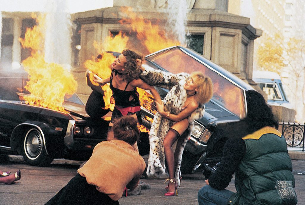 Two scantily clad models fight in the street in front of a burning car as Faye Dunaway as Laura Mars photographs them