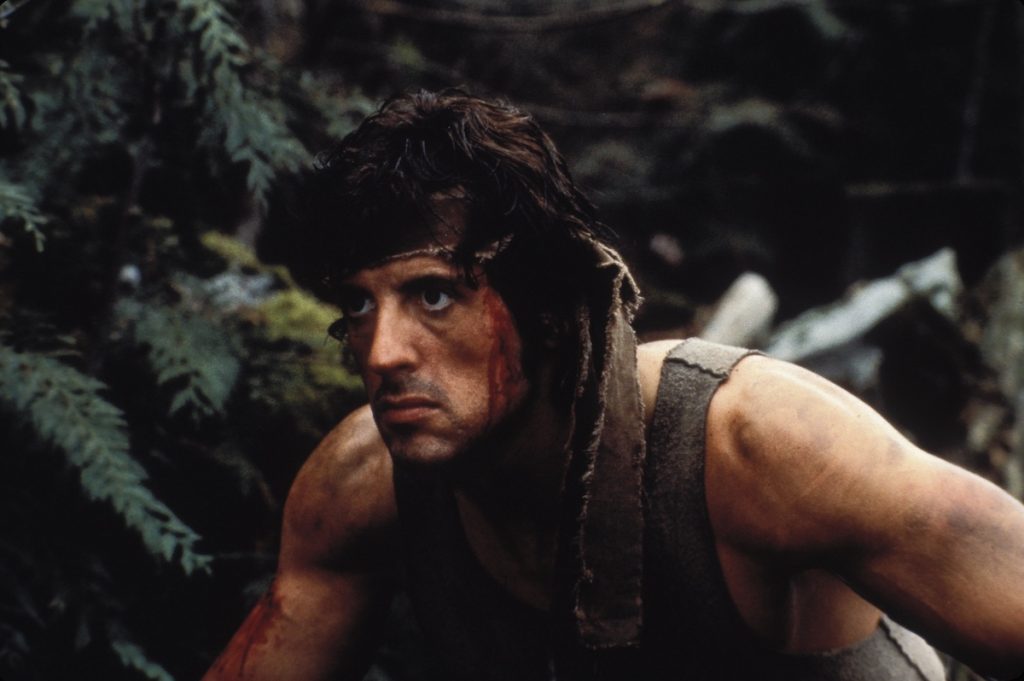 Sylvester Stallone as John Rambo, wears a cloth around his bloodied head, crouches in the woods