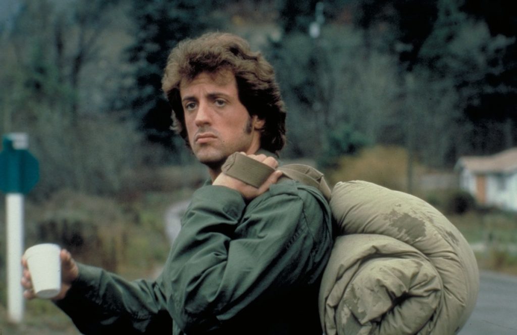 Sylvester Stallone as John Rambo, holds a styrofoam coffee cup in one hand and has a sleeping bag over his shoulder