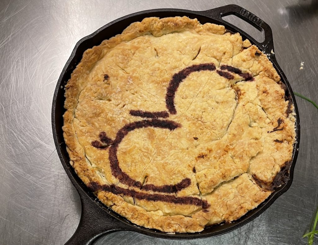 A pot pie in a skillet featuring a brown outline of stylized silhouette of Alfred Hitchock 