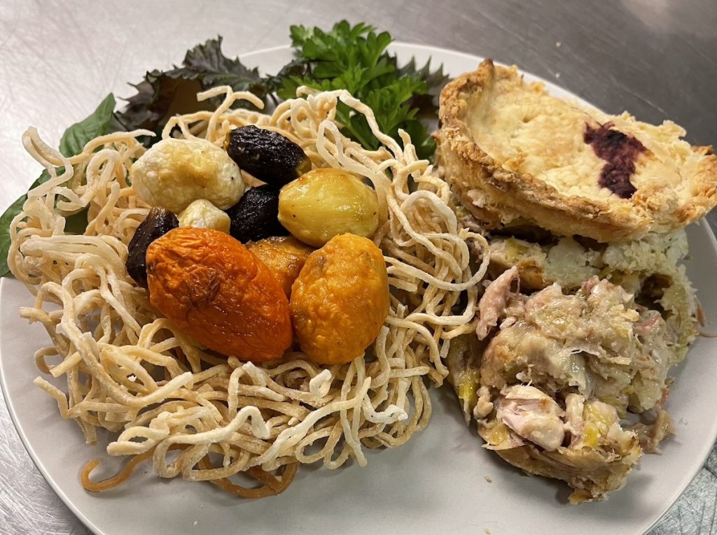 A plate with a serving of pot pie next to a roasted root vegetables carved into eggs served in a nest of fried noodles 