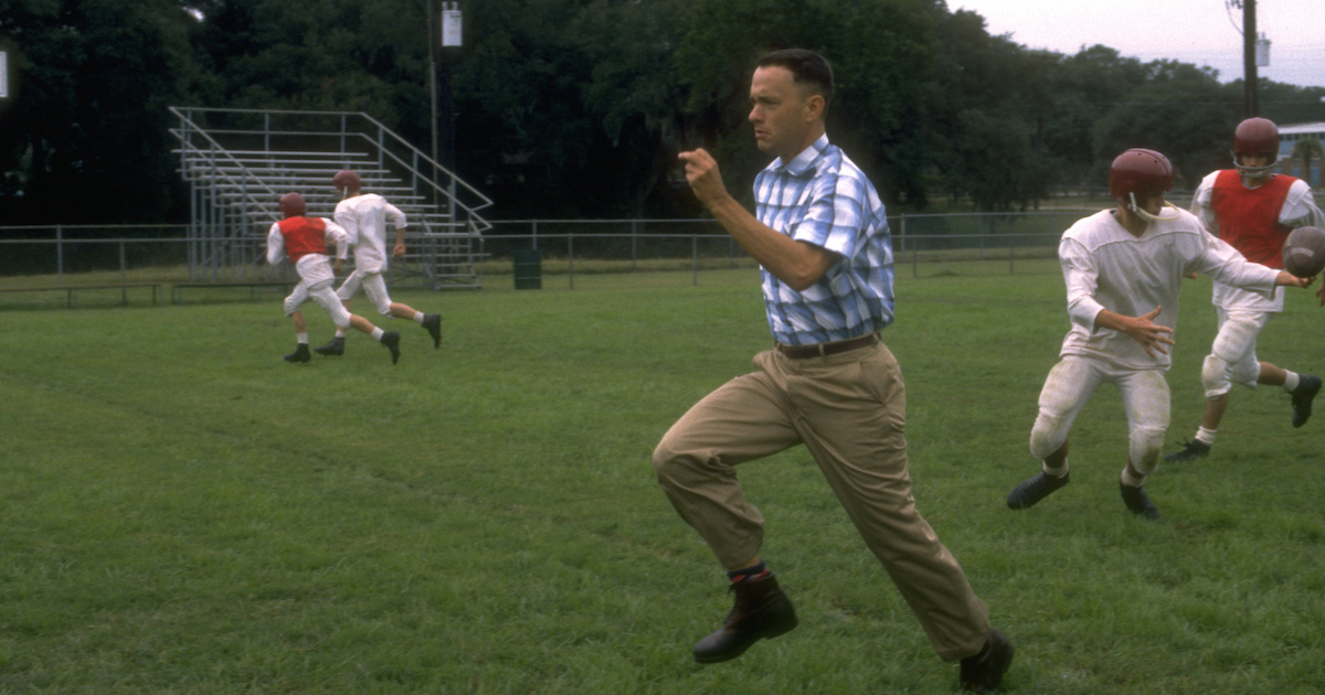 Forrest Gump (Tom Hanks) runs in a field as a football game is played behind him