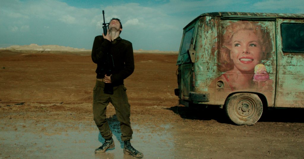A man in a military uniform looks directly up sky as he holds a rifle standing in barren landscape near a van with a faded picture of a woman eating ice cream on the side