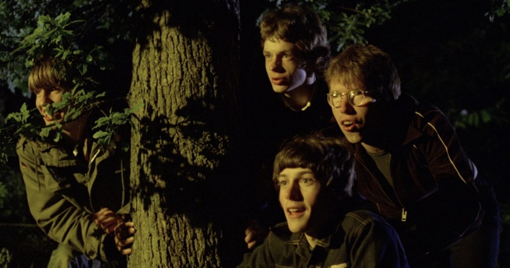 A group of four teen boys peer out excitedly from behind a tree