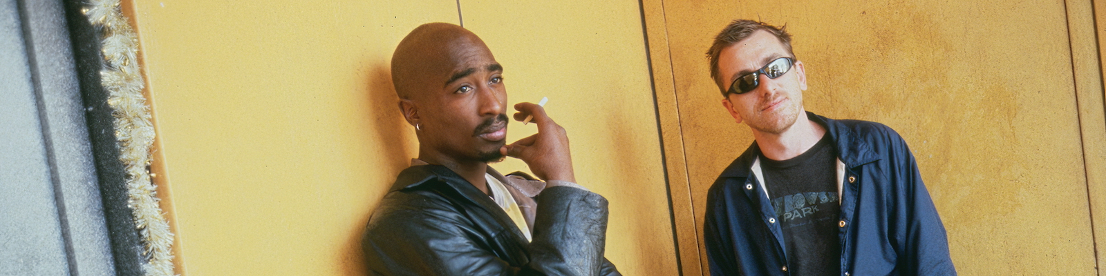 A smoking Tupac Shakur and Tim Roth in sunglasses lean against a yellow wall in an image from the film Gridlock'd