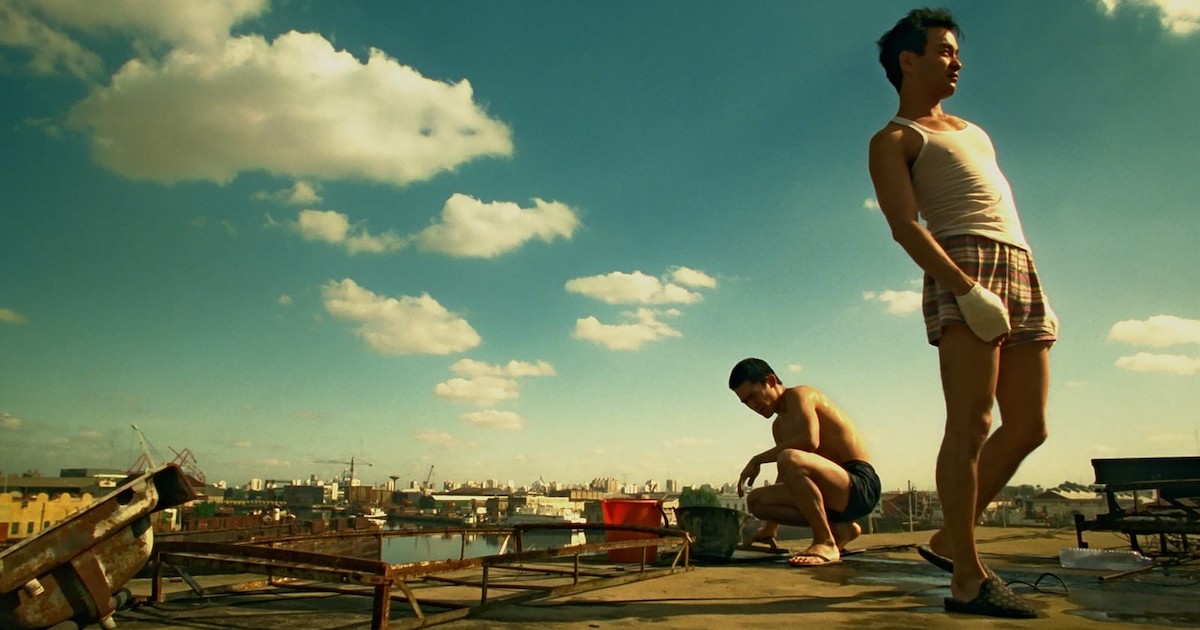 Tony Leung and Leslie Cheung on a urban rooftop in the film Happy together. They are both young Chinese men with slim builds. One stands in the foreground facing right, wearing a white undershirt, boxer shorts and slippers and has a bandage wrapped around his hand. The other crouches in the background facing left, wearing only boxer shorts and flip flops. The rooftop is strewn with rusty scrap metal objects and garbage. The sky is blue with some clouds. Cranes and buildings stretch across the horizon.