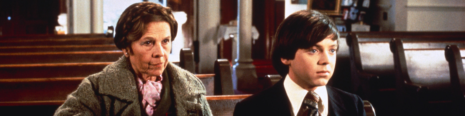 In a church, a white woman in her 80s (Ruth Gordon as Maude) sits in the pew behind and slightle to the right of a white man in his twenties (Bud Cort as Harold). Maude wears her brown hair up in braids. She is in a brown, somewhat dingy wool overcoat with a pink silk scarf peeking out. Harold's dark hair is neatly combed across his head and swoops into his face. He is wearing a black suit coat, white shirt with pointed collars and a brown and silver geometric tie.