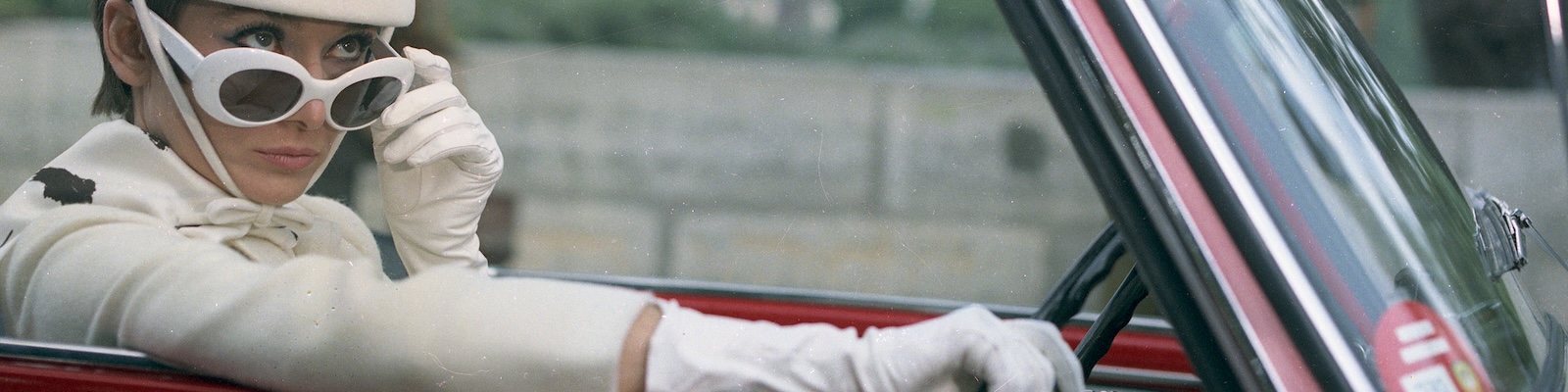 Audrey Hepburn sits in a red convertible wearing all white (coat, gloves, sunglasses, bonnet).