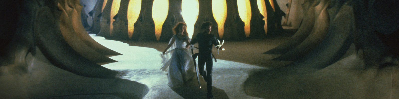 A young man and woman in medieval dress run out of an ominously glowing structure designed to look like it is constructed of bones.