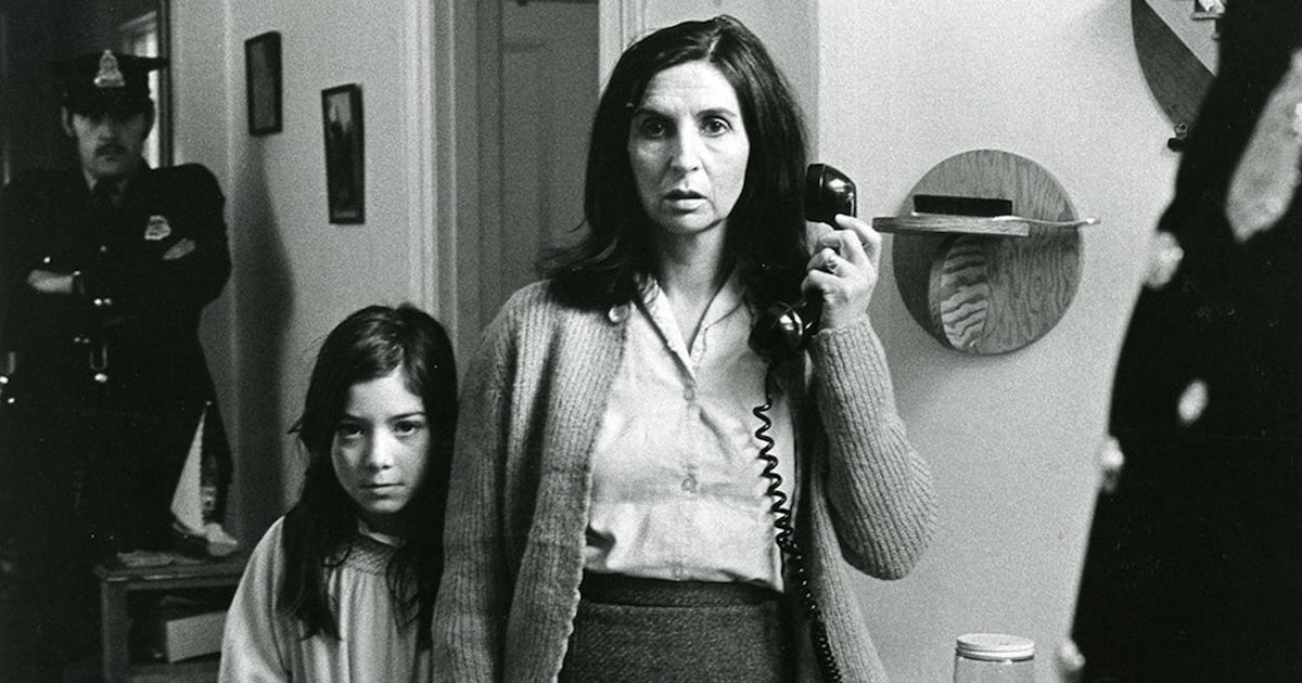 Still photo from the film Les Ordres. A frightened looking white woman around 30 to 40 years of age holds a telephone receiver. Beside her stands a young white girl of around ten. A white uniformed police officer leans on a wall behind them to the left. 