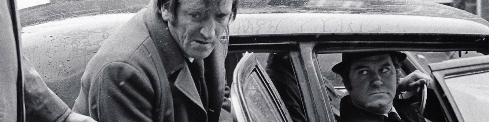 Still photo from the film Les Ordres. A distressed looking middle aged white man is escorted into the back seat of the car as the stern looking white middle aged male driver looks on.