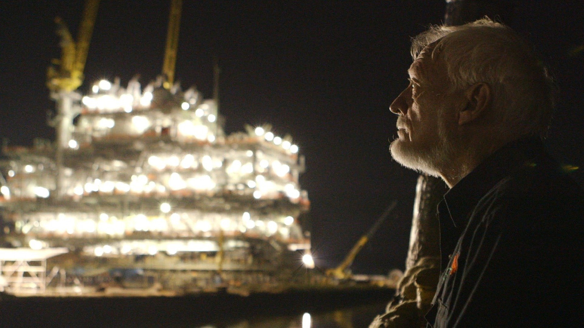 A night a man stands in silhouette in front of the blazing lights of an oil rig