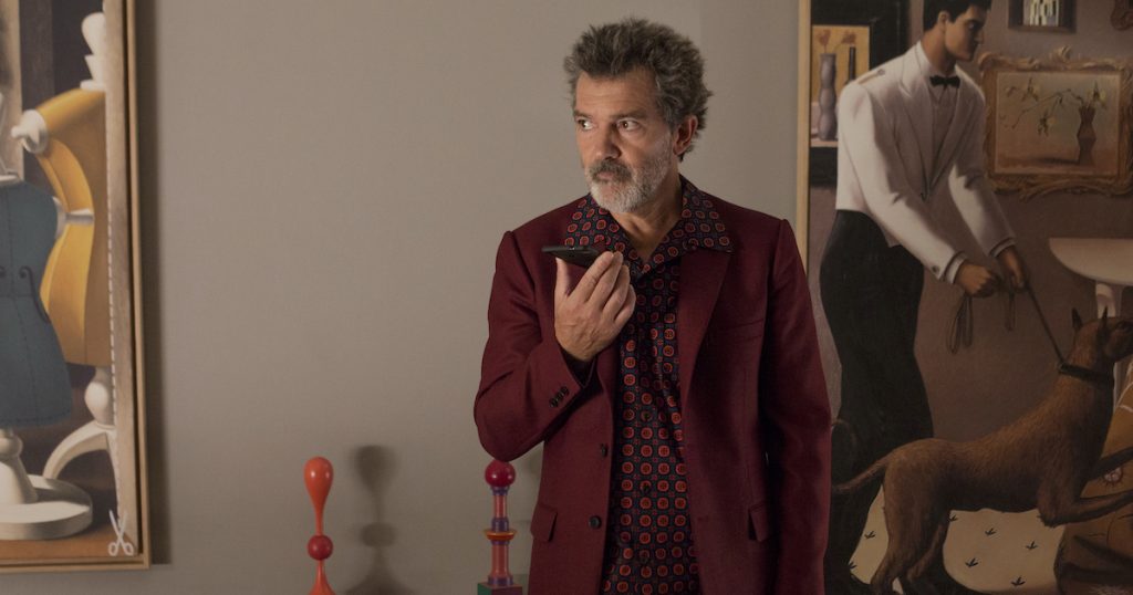 Antonio Banderas in a burgundy suit and wildly patterned shirt speaks into a cell phone in front of a collection of modern art