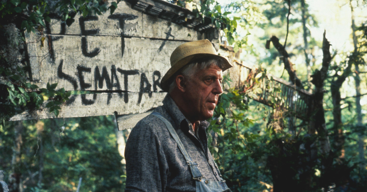An elderly man in denim overalls and a straw hat stands in an overgrown wooded area in front of a handpainted sign reading "Pet Sematary"