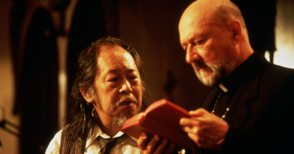 A middle aged Chinese man (Victor Wong) with long scraggly hair and a salt and pepper moustache and chin beard looks at a book held by and middle aged bald white catholic priest with a grey beard. Still photo from the film The Prince of Darkness