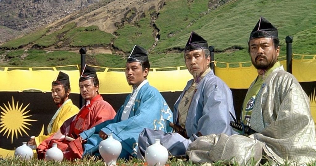 Five men in traditional japanese robes sit in a row in front of a black and yellow banner with a sun emblem. Mountains loom in the distance (Ran)