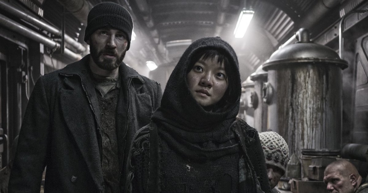 A young bearded white man and a young east asian woman wear ragged winter clothing while standing in a cramped and dirty train car filled with mechanical equipment 