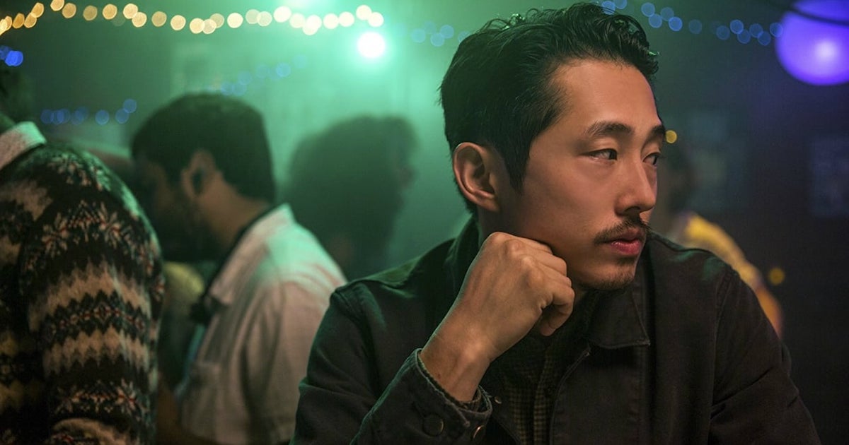 Close up of Steven Yeun looking with his head turned to the side and his fist pressed to his cheek. He is a mid-30s Korean-American man with short wavy black hair and a tidy, closely cropped moustached and goatee. He appears to be in a bar or party behind him several people can be seen standing in a dimly lit room with festive lights strung from the ceiling.