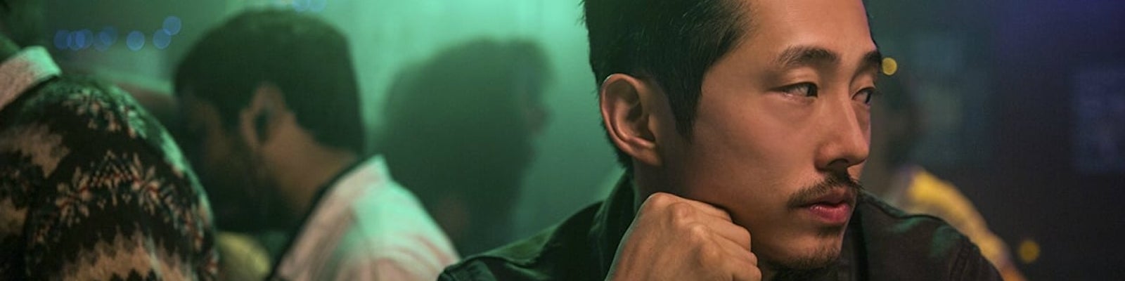 Close up of Steven Yeun looking with his head turned to the side and his fist pressed to his cheek. He is a mid-30s Korean-American man with short wavy black hair and a tidy, closely cropped moustached and goatee. He appears to be in a bar or party behind him several people can be seen standing in a dimly lit room with festive lights strung from the ceiling.