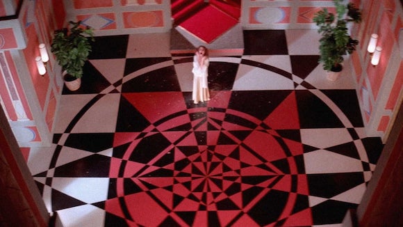 A woman is seen from above screaming while standing in a large ornate room with a striking geometric feautring intersecting lines making triangles on concentric circles floor in red white and black 